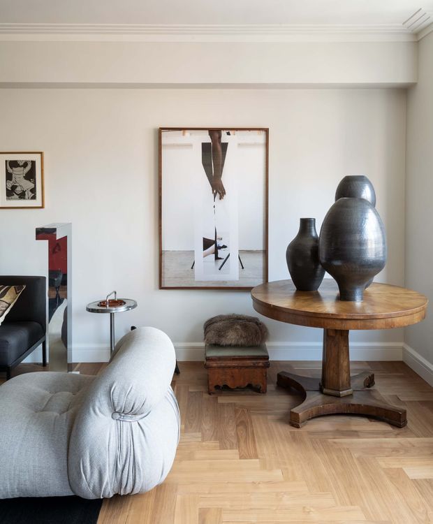 A refined living room with a pale upholstered sofa in the foreground, a round timber pedestal table displaying sculptural dark vessels, and a small upholstered stool. The room has light herringbone timber flooring, warm off-white walls, decorative ceiling cornices, framed artwork, and a dark accent chair and side table. The composition is calm and gallery-like, combining classic architectural detailing with contemporary sculptural furnishings.