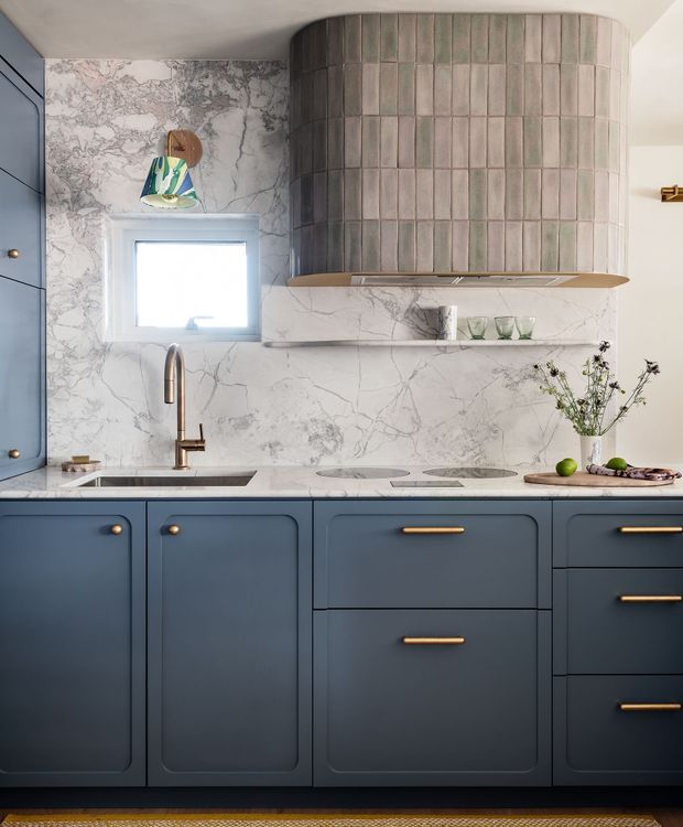 A compact, refined kitchen vignette featuring deep blue shaker-style cabinetry with rounded panel detailing and brass hardware. A pale veined marble-look stone continues across the benchtop and full-height splashback, paired with a dark inset sink and brushed brass tapware. The focal point is a curved, vertically tiled rangehood enclosure in muted taupe, grey and blush tones, with open shelving beneath. A small window, sculptural pendant light, glassware, and minimal produce styling complete the space.