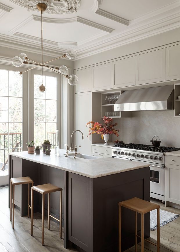 Elegant, light-filled traditional kitchen with a large central island, dark painted base cabinetry, pale upper cabinetry, a substantial range cooker and stainless-steel hood. Tall multi-pane windows and ornate plaster ceiling mouldings add classic architectural character. The island has a pale stone benchtop and seating for several stools, while a sculptural globe pendant provides a contemporary accent.