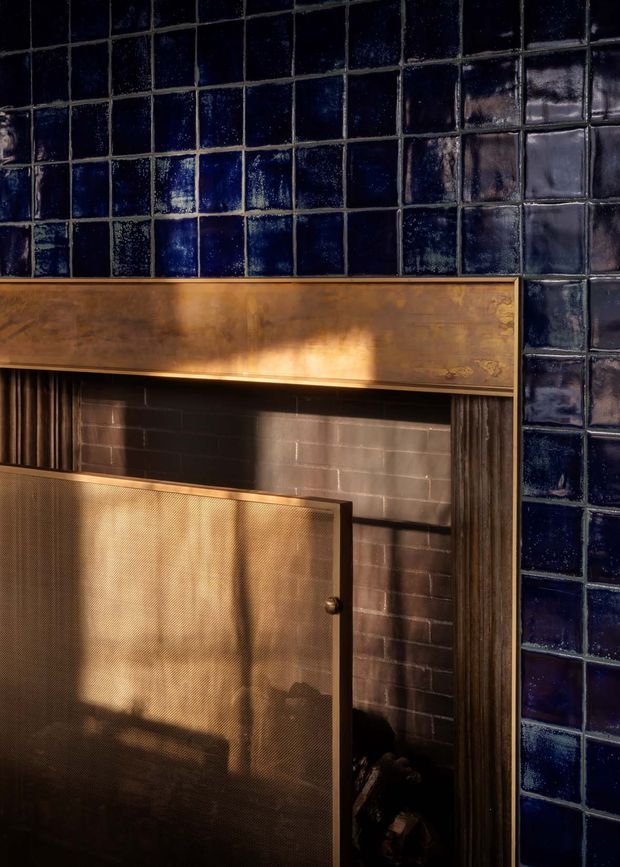 Close-up architectural detail of a traditional fireplace with a brass-toned mantel and perforated metal fire screen, framed by glossy dark navy-blue square tiles. The fireplace surround includes dark masonry and vertical fluted trim, with warm sunlight creating a rich, moody atmosphere.