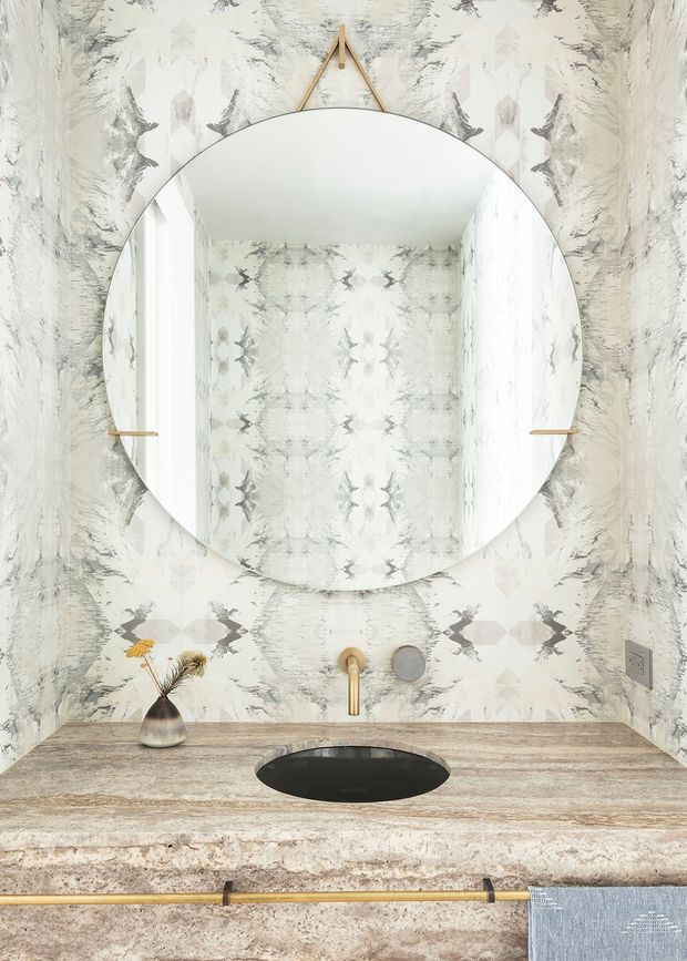 A refined bathroom vanity vignette featuring a large round frameless mirror with a slim brass hanging support, set against expressive grey-and-white patterned wallcovering. The vanity has a thick pale stone benchtop with an integrated black oval basin, brushed brass wall-mounted tapware and hardware, and a small decorative vase. The overall composition is bright, textural, and sophisticated.