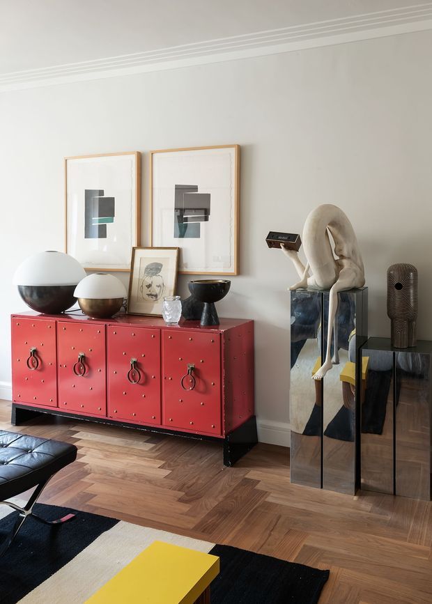 A refined living room vignette with a bold red studded sideboard, sculptural black and white lamps, framed abstract artwork, and a distinctive sculptural figure displayed on mirrored pedestal plinths. The room has pale walls with decorative ceiling moulding, warm geometric timber parquet flooring, and a black-and-cream rug with a yellow coffee table. The overall look is curated, eclectic, and art-focused with strong colour and contrast.