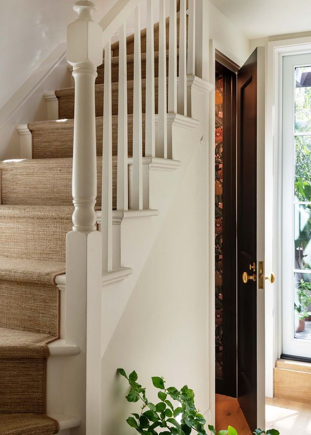 A bright traditional interior focused on a staircase and adjacent entry area. The staircase has a natural woven runner, white painted timber balustrades with turned newel posts, and decorative square-profile trim. Beside it is a tall dark-wood built-in cabinet or closet with brass hardware and patterned interior lining. A white-framed glazed door or window brings in daylight, while a potted green plant sits at the base of the stair.