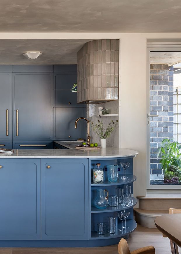 A refined blue kitchen with tall shaker-style cabinetry, long brass handles, a curved island end with open display shelving, and a pale stone benchtop. A curved tiled rangehood enclosure forms a feature architectural element above the cooktop, while a brass tap and integrated sink sit beneath it. The kitchen opens toward a small step-up dining area with a round timber table, and a glazed door reveals an adjacent blue-grey brick wall. The palette is muted blue, pale stone, warm timber and brass, with softly textured plaster ceilings and walls.