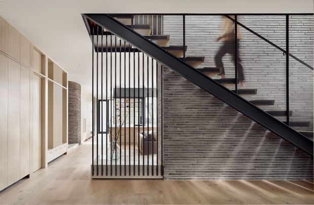 Interior view of a contemporary multi-level home featuring a dramatic black metal staircase with vertical balustrade rods, floating timber treads, and a dark structural stringer. The staircase runs alongside a grey exposed-brick feature wall, with pale timber built-in cabinetry and warm timber flooring. Glazing and an upper-level walkway create a sense of openness and vertical connection.