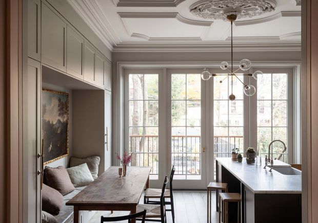 Refined open-plan kitchen and dining area with a built-in banquette, timber dining table, central island with sink, tall French-style windows opening to a balcony, and an ornate coffered ceiling with decorative plaster ceiling medallion. The room combines pale grey cabinetry and trim with dark accents, natural timber furniture, and a sculptural globe chandelier.