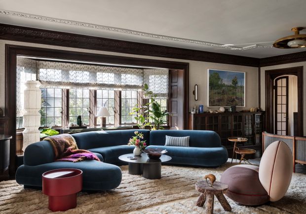 Elegant living room with dark timber wall panelling and trim, ornate ceiling cornice, large multi-pane windows with sheer patterned blinds, and a glazed-panel timber door. A deep blue curved sectional sofa, sculptural pale pink lounge chair, round coffee tables, textured rug, plants, artwork and dark built-in cabinetry create a refined, eclectic interior.