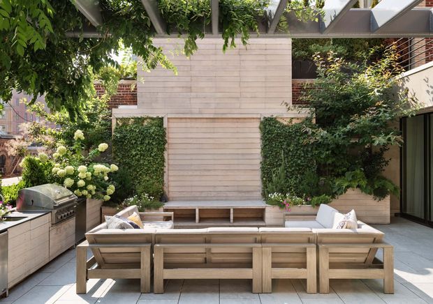 A landscaped contemporary outdoor entertaining area with a built-in outdoor kitchen, barbecue, large timber outdoor sectional seating, tiled paving, and a central timber-clad feature wall with concealed garage-style door. Dense greenery and climbing plants surround the courtyard, while a pergola overhead provides filtered shade.
