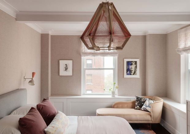 A softly styled bedroom with a bed in the foreground, a blush upholstered sofa beneath the window, built-in white cabinetry, framed artwork, and a distinctive woven geometric pendant light. The room has warm taupe walls, white architectural trims, layered neutral and burgundy textiles, and abundant natural light.