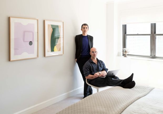 A bright, minimalist bedroom with two framed abstract artworks on a pale wall, a large industrial-style window, and a textured upholstered bed in the foreground. Two people are present, one standing and one seated casually on a chair near the bed. The room has restrained furnishings, white walls, light flooring, and abundant natural light.