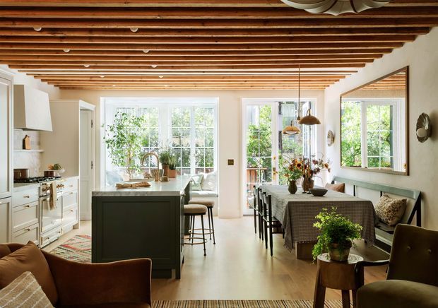 Warm, light-filled open-plan kitchen and dining area with white cabinetry, a dark central island, timber flooring, and a dining table beside large windows. The room features a distinctive exposed timber-beam ceiling, pendant lights, built-in bench seating, and leafy garden views through the glazing. A portion of the living area is visible in the foreground.