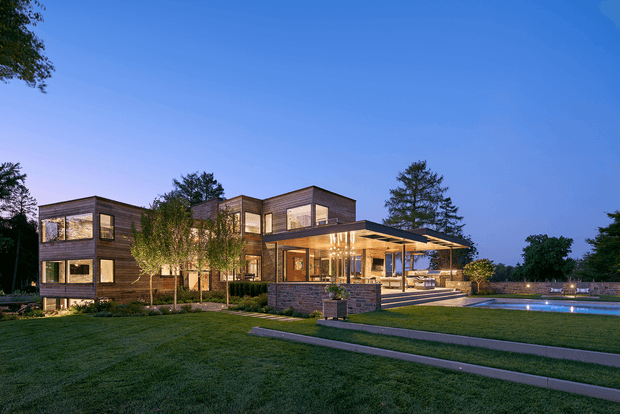Twilight exterior view of a large contemporary multi-storey residence with rectilinear volumes, extensive glazing, dark timber-toned cladding, and a broad covered outdoor entertaining terrace. The landscaped lawn, paved walkway, low masonry retaining walls, and illuminated swimming pool are clearly visible.