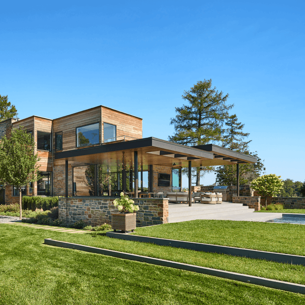 A contemporary multi-level residence viewed from the landscaped garden, featuring warm horizontal timber cladding, extensive floor-to-ceiling glazing, a broad flat-roofed covered outdoor entertaining area, and a low stone-clad retaining wall. The property has manicured lawns, stepped garden edging, outdoor seating, and a swimming pool visible at the right edge.