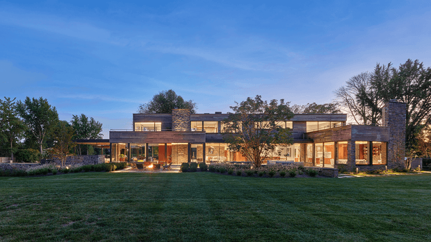 Wide dusk exterior view of a large contemporary two-storey residence set behind an expansive lawn. The house has a low, horizontally composed form with extensive floor-to-ceiling glazing, warm interior lighting, flat rooflines, dark stone-clad vertical volumes, timber-toned soffits or screens, and landscaped planting along the facade.