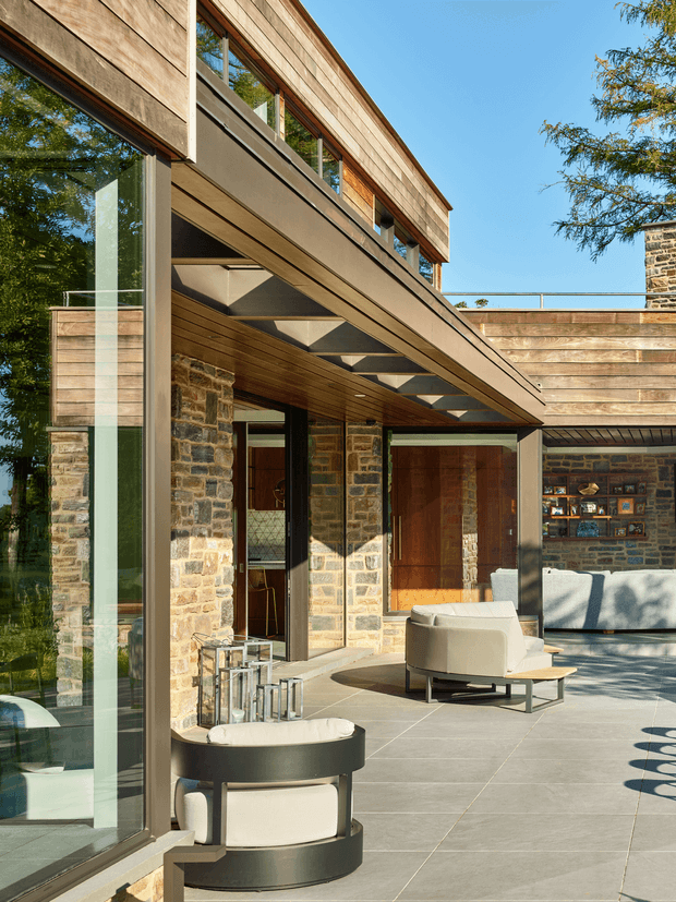 A sunlit contemporary courtyard terrace adjoining a stone-clad house. Large floor-to-ceiling glazing, warm timber soffits and horizontal timber cladding frame an open outdoor living area with pale upholstered lounge chairs, a sculptural round side table, and a built-in stone fireplace wall. A steel-framed cantilevered upper level projects over the terrace, with visible structural beams and a glass balustrade. The composition combines natural stone, timber, dark metal, and expansive glazing in a refined indoor-outdoor setting.