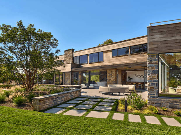 Exterior view of a large contemporary two-storey residence with extensive glazing, horizontal timber cladding, and rugged stacked-stone walls. The home opens onto a landscaped courtyard and outdoor entertaining terrace, with stepping-stone paving through lawn and ornamental planting.