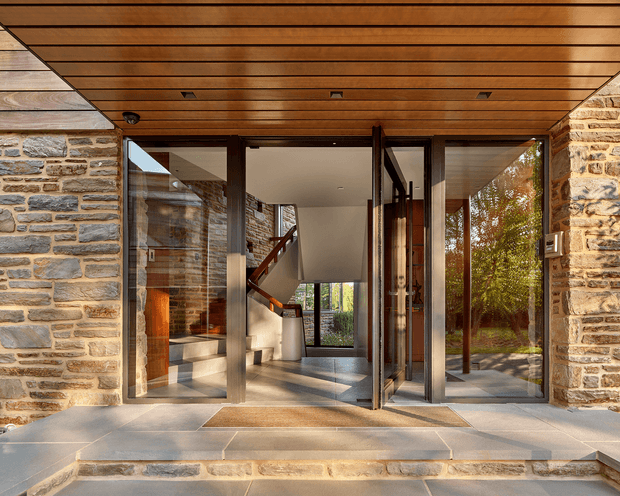 Front entry of a contemporary stone-clad residence, viewed from outside beneath a warm timber soffit. Large dark-framed glazed doors open into a bright foyer with a visible staircase, polished light flooring, and timber detailing. Natural stone walls and extensive glazing connect the entry to the surrounding landscape.