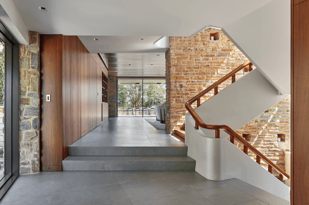 A contemporary residential entry hall and stairway with broad grey stone or porcelain tiled flooring, warm timber wall panelling and cabinetry, exposed stacked-stone walls, and a sculptural staircase with a polished timber handrail. The space opens toward full-height glazing and a bright outdoor outlook, with a linear slatted ceiling detail extending through the hall.