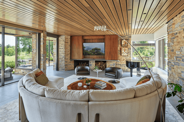 A spacious, refined living room with a large curved cream sectional arranged around a round timber coffee table. The room features exposed natural stone walls, extensive floor-to-ceiling glazing, a warm slatted timber ceiling, built-in timber media cabinetry, leather lounge chairs, and a sculptural arched floor lamp. Outdoor greenery and landscape are visible through the glazing.