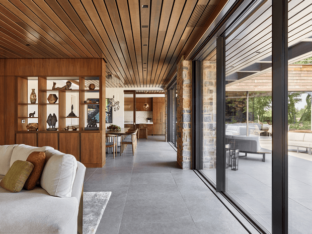 A refined open-plan residential interior with a living area in the foreground, dining area and kitchen beyond, and extensive black-framed sliding glazing opening to a covered outdoor terrace. The space features a warm timber-lined ceiling and built-in timber display joinery, exposed natural stone wall sections, large-format grey floor tiles, and a neutral upholstered sofa with earthy cushions. The indoor-outdoor connection and landscaped terrace are clearly visible through the glazing.