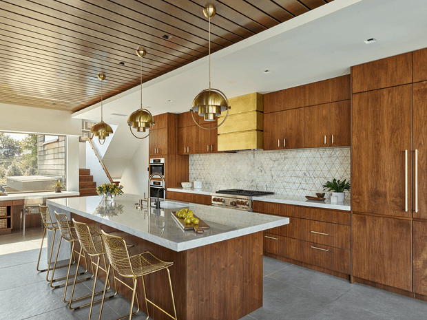 A spacious contemporary kitchen with extensive warm timber cabinetry, a large stone island with integrated seating, and a geometric tiled splashback. Brass pendant lights hang from a slatted timber ceiling, while large glazing brings in abundant daylight. The open-plan space includes a dining or living area visible to the left and a stairway in the background.