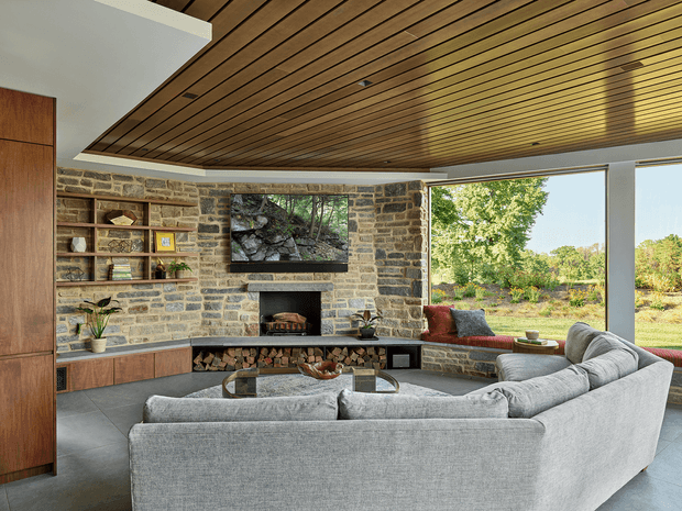 A spacious contemporary living room with a large grey sectional sofa facing a stone-clad fireplace and television wall. Built-in timber cabinetry and open shelving flank the fireplace, while broad glazed openings provide views to a wooded landscape. The room features a striking slatted timber ceiling, large-format grey flooring, and an integrated window seat with cushions.