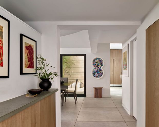 A bright, refined residential interior viewed from a hallway or entry passage. The foreground contains a fixed timber-fronted console or sideboard with a dark stone benchtop, while framed artwork and a potted plant decorate the wall. Beyond is a dining area with a timber table and chairs, large-format pale floor tiles, a dark-framed glazed opening, and light timber built-in cabinetry. The space has clean white walls and ceilings with contemporary detailing.