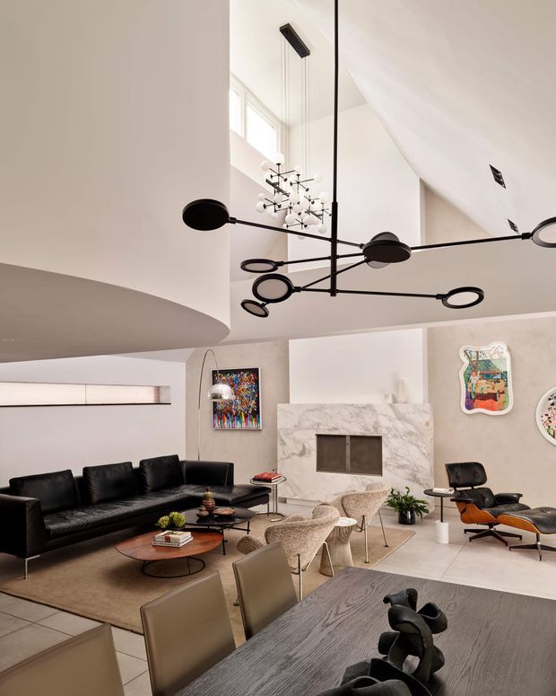 A sophisticated open-plan living and dining area with a dramatic double-height ceiling. The room features a black leather sofa, sculptural lounge chairs, a marble fireplace surround, contemporary artwork, a large dining table with upholstered chairs, and striking modern pendant lighting. The neutral palette of white, beige, grey, and black creates a refined, gallery-like atmosphere.