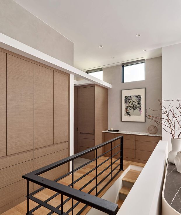 A bright modern upper-level landing overlooking a stairwell, with extensive built-in warm wood cabinetry, a black metal horizontal-bar balustrade, white plaster walls and ceiling, and high clerestory windows. A timber staircase descends beside a compact built-in wood console, with minimalist artwork and neutral decorative objects adding understated styling.