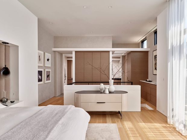 A bright, refined bedroom with a partially screened dressing and storage area. The room features warm timber flooring, soft white bedding, pale built-in cabinetry, a central low dresser or island, timber wall panelling, framed artwork, and full-height sheer curtains. Recessed ceiling lighting and abundant daylight create an airy, contemporary interior.