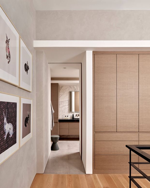A refined contemporary interior view looking from a timber-floored room through a narrow hallway into a bathroom or dressing area. The space features full-height light timber built-in cabinetry, pale textured plaster walls, white structural beams, warm timber flooring, and a dark-framed console or table in the foreground. Framed artwork lines the left wall, while the rear room includes a dark vanity benchtop, timber cabinetry, mirror, and pale stone or tile surfaces.