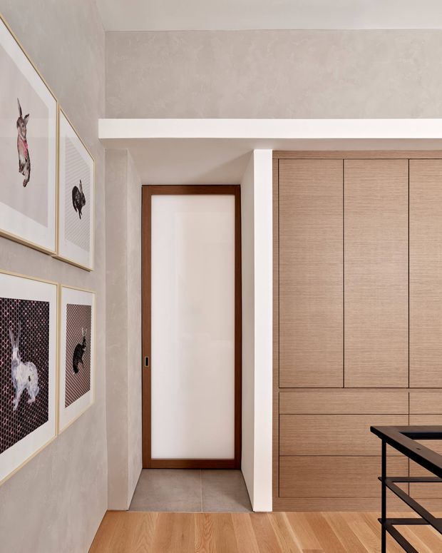A refined interior hallway or living area with warm light timber flooring, pale textured plaster walls, a dark timber-framed frosted glass door, and extensive built-in light timber cabinetry. Framed artwork is arranged along the left wall, while part of a black metal-framed counter or stair railing appears at the lower right. The space has clean contemporary detailing and a warm neutral palette.