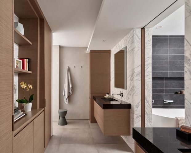 A refined contemporary bathroom with a compact open vanity, wall-mounted timber-toned cabinetry, black stone benchtop, and integrated basin. The space includes a toilet and towel rail at the rear, full-height marble-look wall panels around the vanity, and a bathtub or shower area finished in dark horizontal tiles. Built-in timber shelving with books and decorative objects occupies the left side. The palette is warm neutral with dark contrasting surfaces and clean-lined detailing.