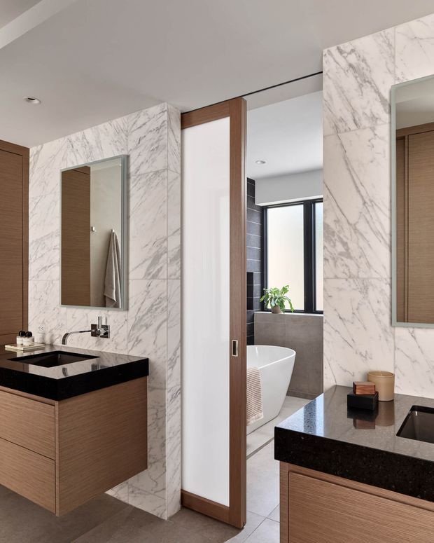 Contemporary bathroom with dual floating timber-veneer vanities, black stone benchtops and integrated basins, large white marble-look wall panels, frameless mirrors, and a partially open timber-framed door revealing a freestanding bathtub and separate bathing area. The space has clean lines, recessed lighting, warm wood accents, and a refined neutral palette.