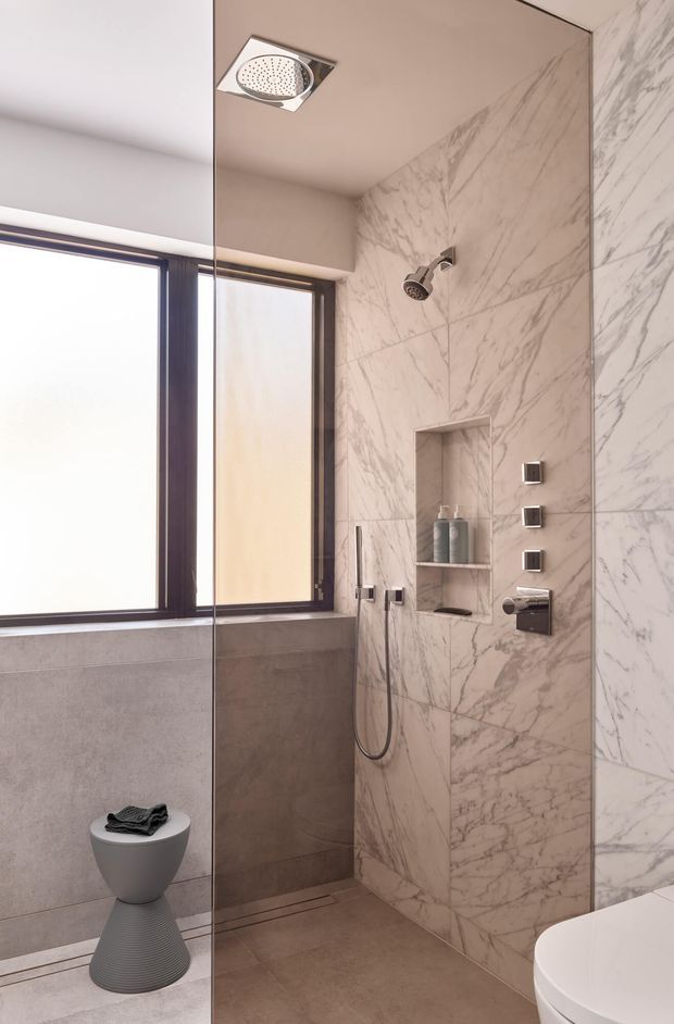 Contemporary bathroom featuring a walk-in shower clad in large-format white marble-look tiles with grey veining, a built-in recessed niche, ceiling and wall-mounted shower fittings, dark-framed frosted windows, and warm taupe-grey floor and lower wall tiles. A wall-hung toilet and sculptural grey stool are visible.