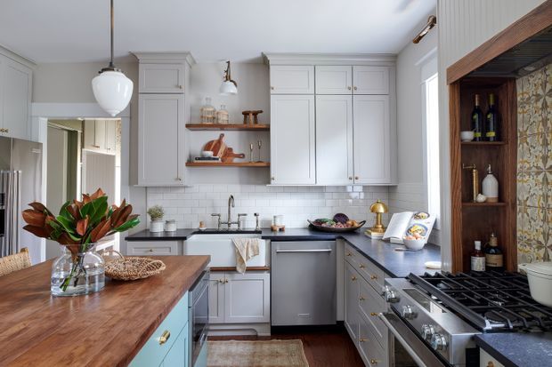 A bright, traditionally detailed kitchen with white Shaker-style cabinetry, dark countertops, a central sink, open timber shelving, and a large stainless-steel range. A warm timber island with blue-green cabinetry occupies the foreground, while patterned tile and dark timber framing create a feature cooking zone on the right. Decorative pendant and wall lighting, brass hardware, and natural wood accents add warmth.