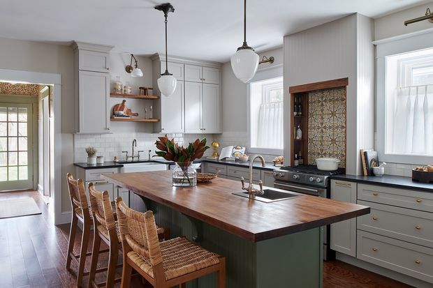 A bright, spacious traditional kitchen with a large timber island and integrated seating for four. The room features pale grey shaker cabinetry, dark stone or composite benchtops, white subway tile splashbacks, timber flooring, brass hardware and tapware, pendant lighting, and a freestanding range cooker. A decorative patterned tile feature is framed beside the cooker, while tall windows and glazed doors provide ample natural light.