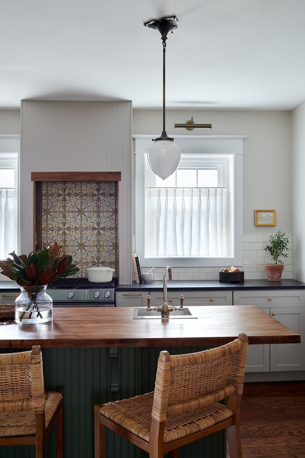 A bright, refined kitchen with a large timber island and woven rattan bar chairs. The space features pale grey shaker-style cabinetry, a dark benchtop, white subway-tile splashback, a decorative patterned tile feature behind the freestanding range, white-trimmed windows with sheer curtains, timber flooring, and a traditional pendant light. Brass and dark metal hardware add contrast, while greenery and a small framed artwork provide subtle accents.