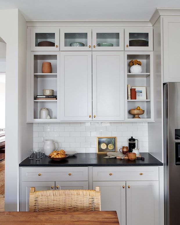 A bright, refined kitchen cabinetry wall with pale grey shaker-style built-in cupboards, glass-front upper cabinets, open display shelving, brass knobs, and a dark stone benchtop. A white subway-tile splashback runs behind the counter, with styled ceramics and glassware on the shelves. A woven timber chair and warm timber island surface appear in the foreground, while a stainless-steel refrigerator is visible at right.