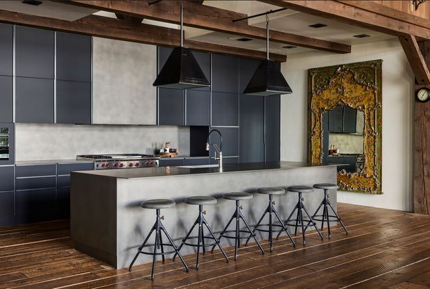 A spacious industrial-style kitchen with a large dark island and integrated sink, six black metal stools, dark flat-panel cabinetry, a stainless steel cooking range, and twin black pendant lights. Exposed timber beams and timber flooring add warmth, while a large ornate framed mirror is mounted on the wall.