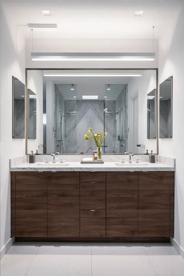 A bright, refined contemporary bathroom featuring a wide double vanity with dark wood-grain cabinetry, a white veined stone benchtop, twin basins and wall-mounted taps. A large framed mirror spans the vanity, reflecting a spacious glass-enclosed marble-look shower. Recessed ceiling lighting and linear vanity lighting create an even, clean illumination.