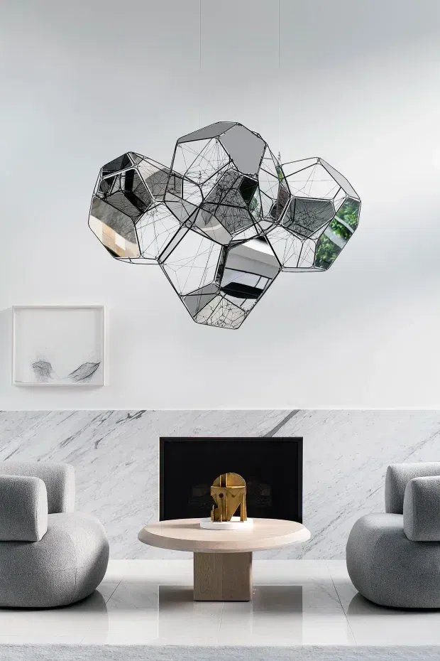 A refined contemporary living room vignette with two sculptural grey lounge chairs facing a small round pedestal coffee table. A black-framed fireplace with a gold sculptural object sits against a dramatic white veined marble feature wall, while a large geometric mirrored wall sculpture dominates the upper wall. The room has a pale, monochromatic palette with polished light flooring and minimalist artwork.