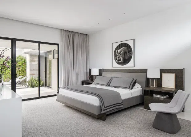 A bright, refined contemporary bedroom with a large upholstered bed, grey layered bedding, dark wood bedside cabinetry, matching table lamps, and a sculptural grey lounge chair. Full-height sliding glass doors with sheer curtains open to an outdoor area, while a large pale carpet and white walls create an airy neutral setting.