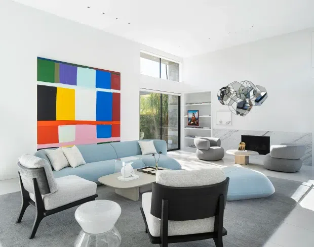 Bright, spacious contemporary living room with a pale blue sectional sofa, sculptural lounge chairs, a large grey rug, and a low central coffee table. White walls and ceiling, expansive floor-to-ceiling glazing, built-in shelving, and a bold geometric multicoloured artwork create a clean modern interior. A sculptural faceted wall light or art piece provides a distinctive focal point.