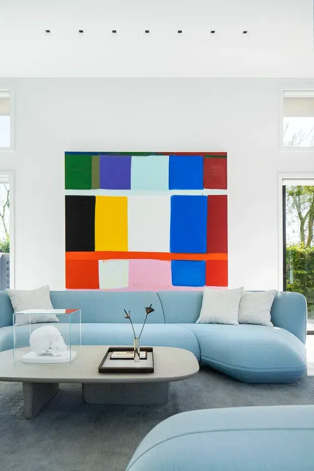 Bright, airy living room with a large light-blue curved sectional sofa, pale rounded coffee table, neutral carpet, and white walls. A bold abstract multicolour artwork forms the main focal point above the sofa. Tall windows and small clerestory-style openings bring in abundant natural light, with a minimalist linear ceiling light detail.
