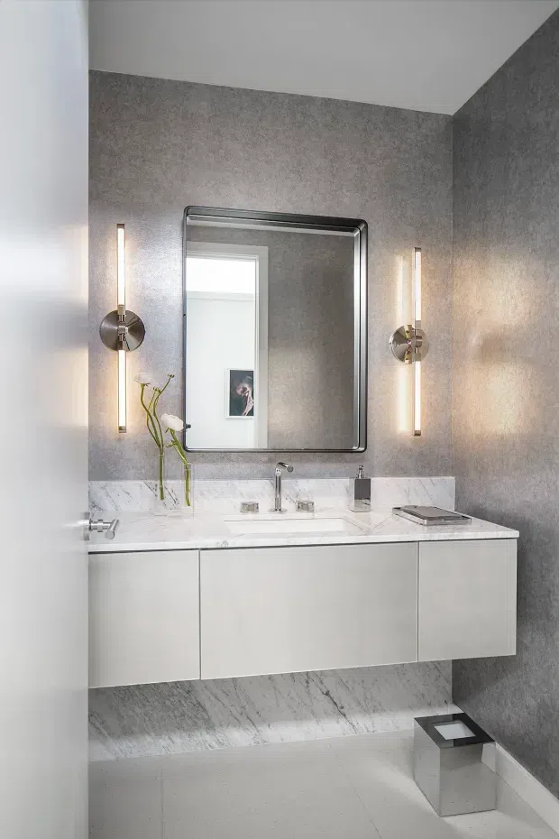 A refined contemporary bathroom vanity with a floating white cabinet, white marble benchtop and matching marble wall wainscot, grey textured wall finish, large framed mirror, and symmetrical vertical wall sconces. The room has a clean, polished, monochromatic appearance with chrome tapware and minimal accessories.