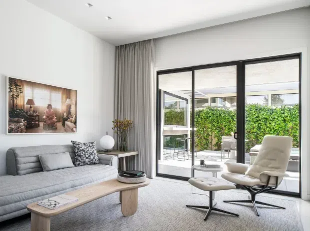 Bright, refined contemporary living room with a grey upholstered sofa, light timber oval coffee table, sculptural lounge chair and ottoman, and a large black-framed glazed opening to a landscaped outdoor courtyard. Neutral furnishings, floor-to-ceiling curtains, recessed ceiling lights, and a framed artwork create a calm, polished interior.