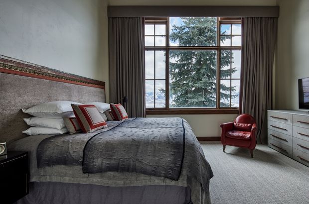 A refined bedroom with a large upholstered bed, layered grey bedding, red accent cushions, and a broad multi-pane timber-framed window overlooking a tree. Full-height dark curtains frame the window, while a red leather armchair and pale built-in chest of drawers sit on a light carpet. Decorative horizontal wall trim adds a traditional architectural detail.