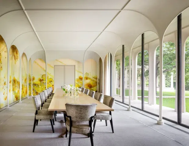 A long, bright dining room with a large rectangular timber dining table surrounded by upholstered chairs. The space features a curved white vaulted ceiling with segmented panels, repeated arched openings, floor-to-ceiling glazing, pale carpet, and a vivid yellow abstract mural along the left wall. Lush greenery is visible outside through the windows.