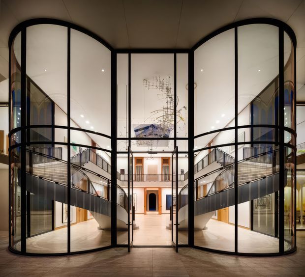 A dramatic interior atrium with two large curved, black-framed glazed volumes flanking a central passage. The space features pale polished flooring, multi-level circulation galleries with black metal balustrades, high ceilings, and a symmetrical view toward a more traditional-looking interior facade with timber-framed doors and artwork. The design combines contemporary glazing and metalwork with an open, gallery-like communal space.